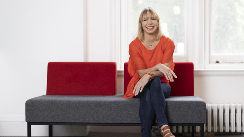 IMechE Chief Executive Alice Bunn sits in front of a window on a grey sofa with red cushions. 